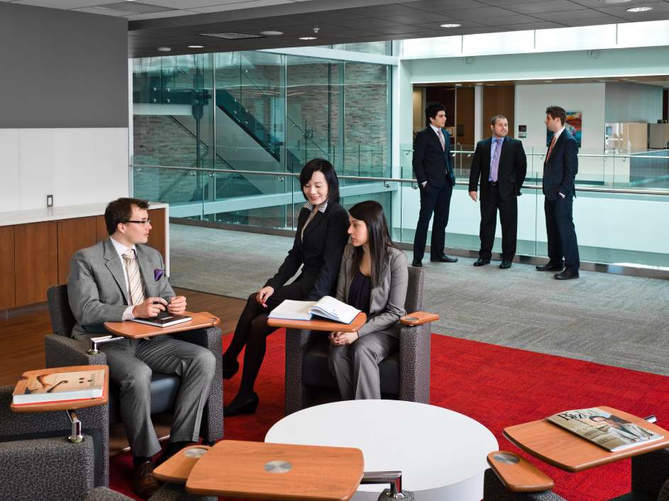 Several businesspeople in a modern lobby, three seated in armchairs with open notebooks, and three standing near the glass railing, conversing.