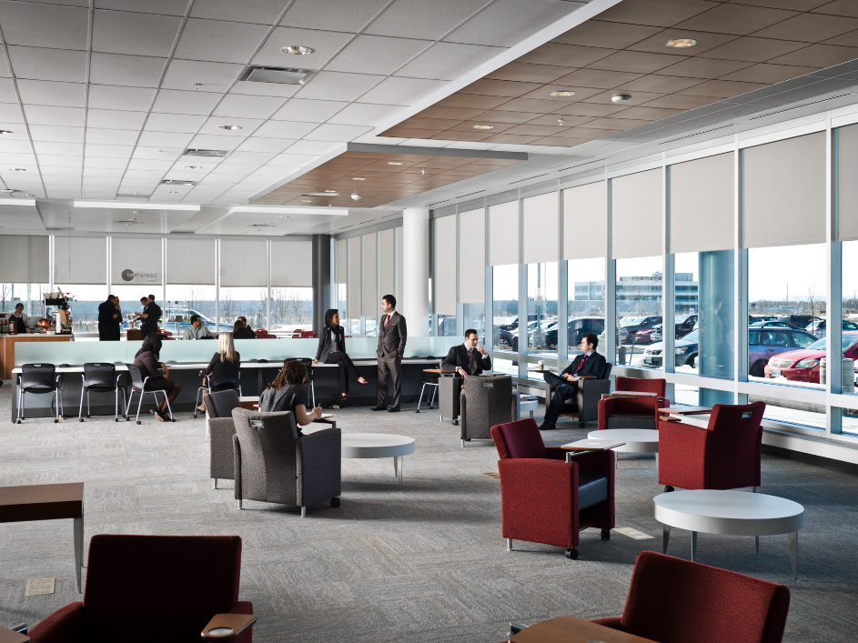 Several businesspeople in a modern lobby with large windows and grey carpet, some seated studying and others standing conversing.